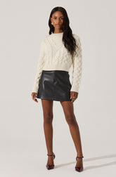 A woman is wearing a cream-colored, cable-knit sweater with a high neckline, paired with a black leather mini skirt. She has on dark brown high heels with ankle straps and gold jewelry, including earrings and rings.